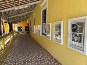 #PraCegoVer Fotografia da varanda da Casa Amarela Eusélio Oliveira, do lado direito estão vários quadros na parede. A casa tem paredes amarelas e um piso marrom e branco, na foto é possível ver ainda parte do telhado e janelas.